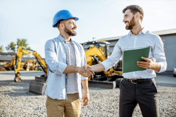 Constructor eligiendo maquinaria pesada para la construcción con un asesor de ventas dándose la mano en el descampado de una tienda con vehículos especiales.