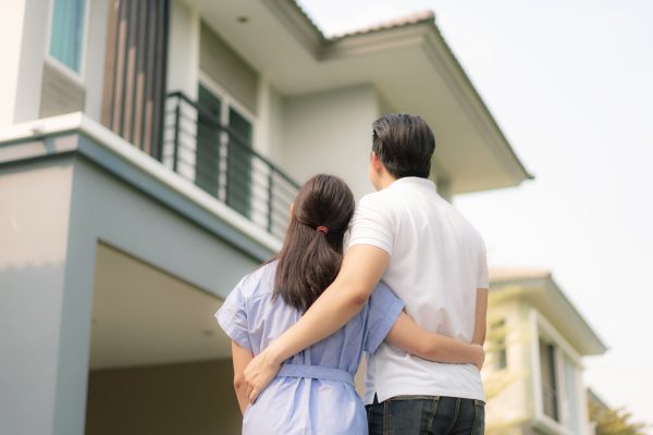 Retrato de espalda de joven pareja asiática de pie y abrazándose que parecen felices delante de su nueva casa para empezar una nueva vida. Concepto de familia, edad, hogar, bienes inmuebles y personas.