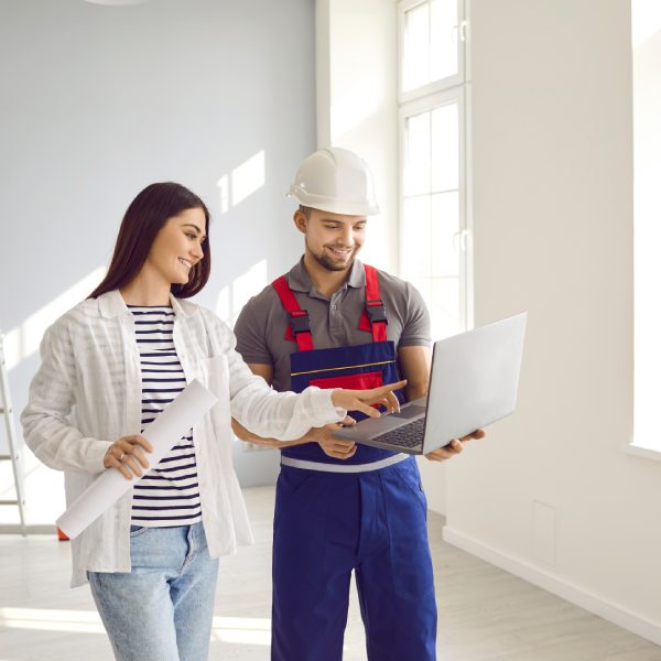 Woman discusses future house building with architect or engineer. Happy young girl with paper roll and man builder in safety hat and uniform look at construction plan on modern laptop computer device