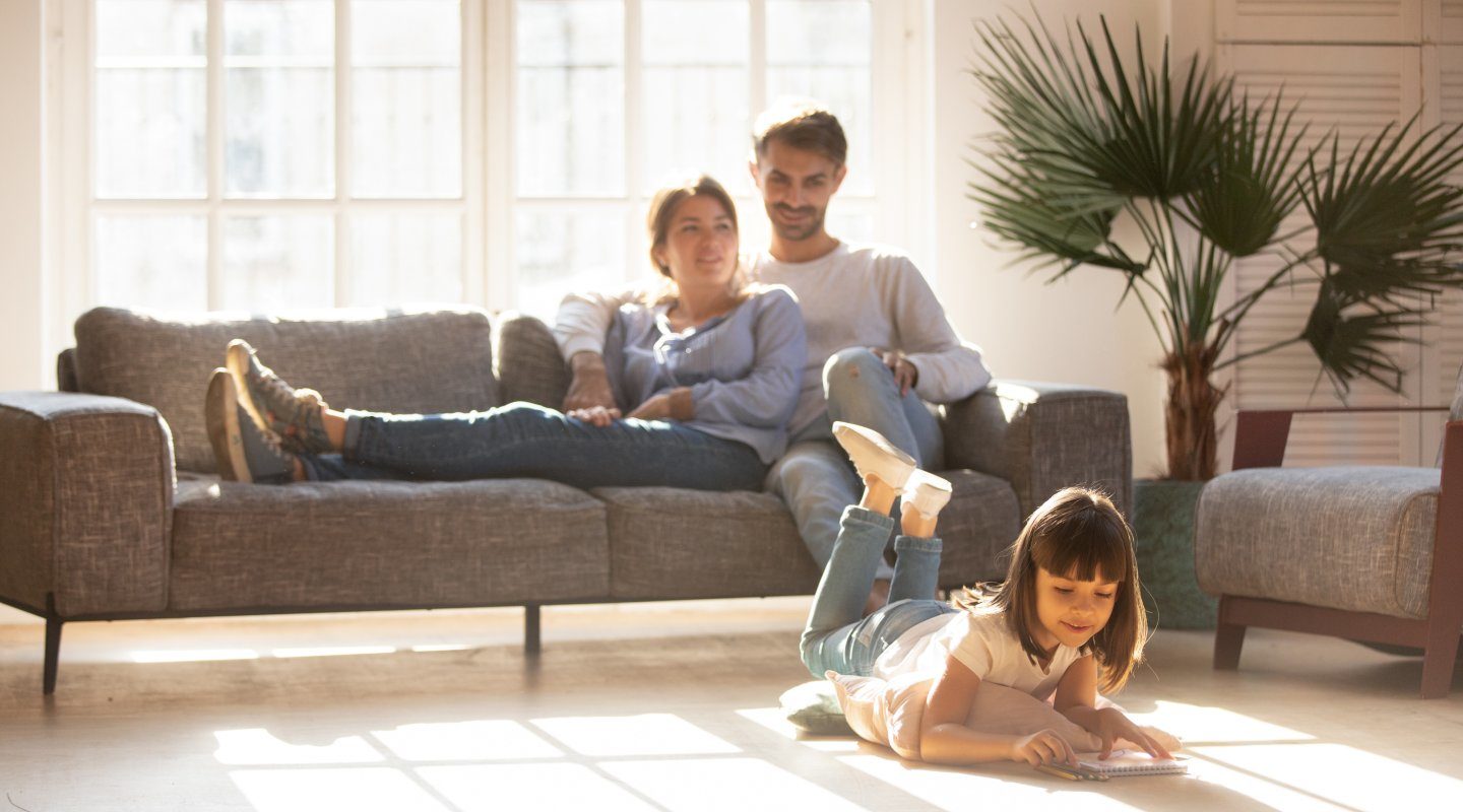 Happy parents relaxing on couch in comfort light living room while little kid child daughter playing on warm floor drawing with colored pencils, family having fun together, underfloor heating concept