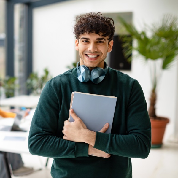 Man college student holding books and smiling at camera while female classmates studying with laptop on the background, coworking space