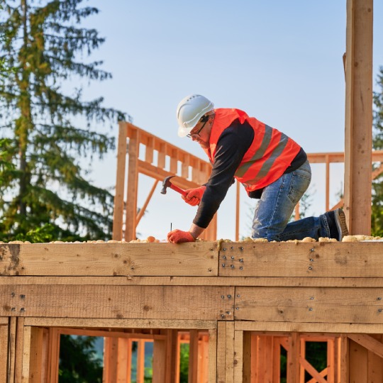 Carpenter constructing two-story wooden frame house near forest. Bearded man hammering nails into the structure, wearing protective helmet, construction vest. Concept of modern ecological construction