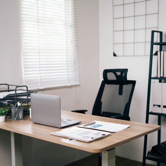 A clean and organized office with a desk, chair, and bookshelf. The desk has a laptop and a stack of papers. The room is well-lit and has a professional atmosphere