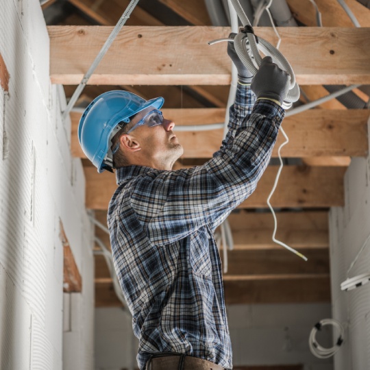 Caucasian Professional Electrician in His 40s Installing Ceiling Light Point Inside Newly Developed Concrete Brick House with Wooden Beams Roof Structure.