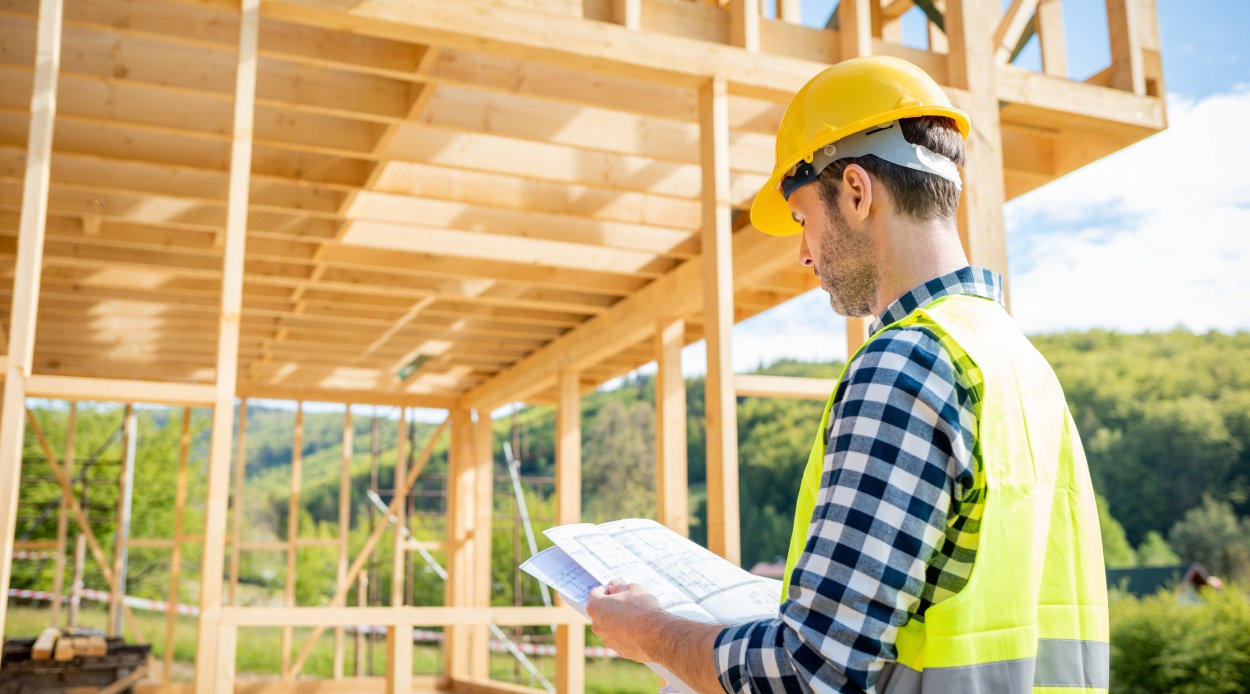 Engineer with hardhat and blueprints on building site of wood frame house under construction