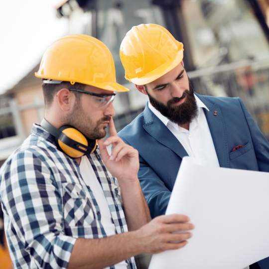 Two engineer builders with blueprint plan at construction site.