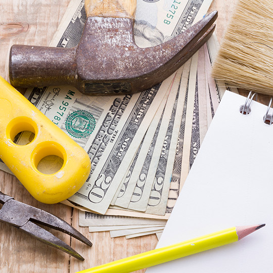 Tools for commercial construction are arranged on a wooden table, including a hammer, pencil, notepad, and paint supplies.