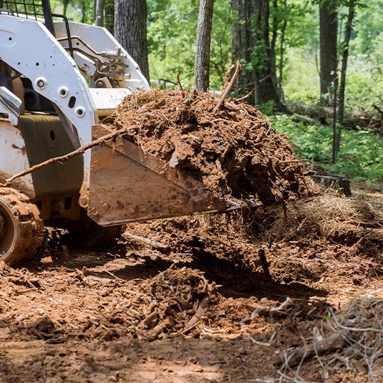 A white skid steer loader operates in a wooded area, focusing on excavation for flood-prone regions in McAllen.