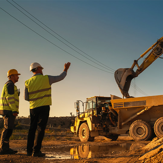 Two men wearing yellow vests are next to a dump truck, involved in excavation efforts in McAllen's flood-prone regions.