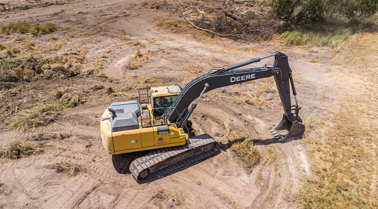 A yellow excavator is actively digging in a dirt field, representing land clearing for construction purposes.