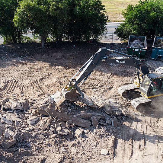 An excavator is actively demolishing a residential structure on a construction site, with rubble scattered around.