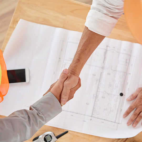 Two people engaged in a handshake above a blueprint, representing collaboration in the commercial construction industry.