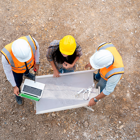 three men wearing hard hats and safety vests gather around a table with a laptop, reviewing custom home building costs in McAllen, TX.